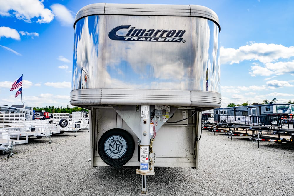 Shiny Cimarron Trailers aluminum trailer parked in a gravel lot with other trailers and a flag in the background.