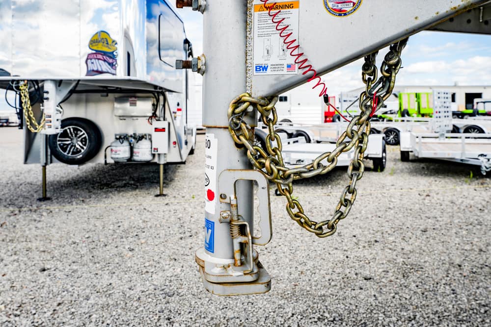 Close-up of a trailer hitch with safety chains on a gravel lot, parked RVs visible in the background.