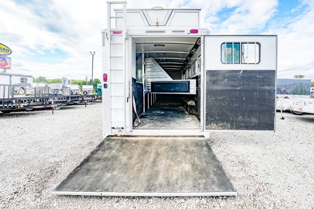 White horse trailer with its rear ramp lowered, showing the empty, dark interior and divider, parked on a gravel lot with other trailers nearby.