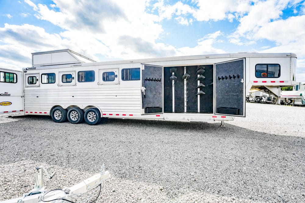 White horse trailer with its side doors open to reveal a tack room, showing saddles hanging on racks and bridle hooks along grey panels in a well-organized interior.