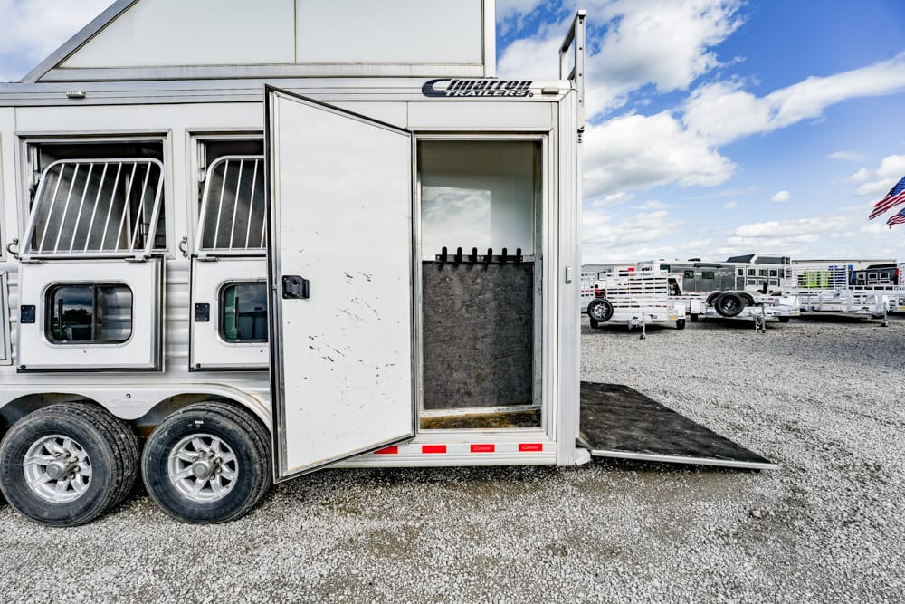 Horse trailer with its rear door open and a lowered ramp in a gravel lot; inside shows a tack area and stalls, with other trailers in the background and an American flag farther away.