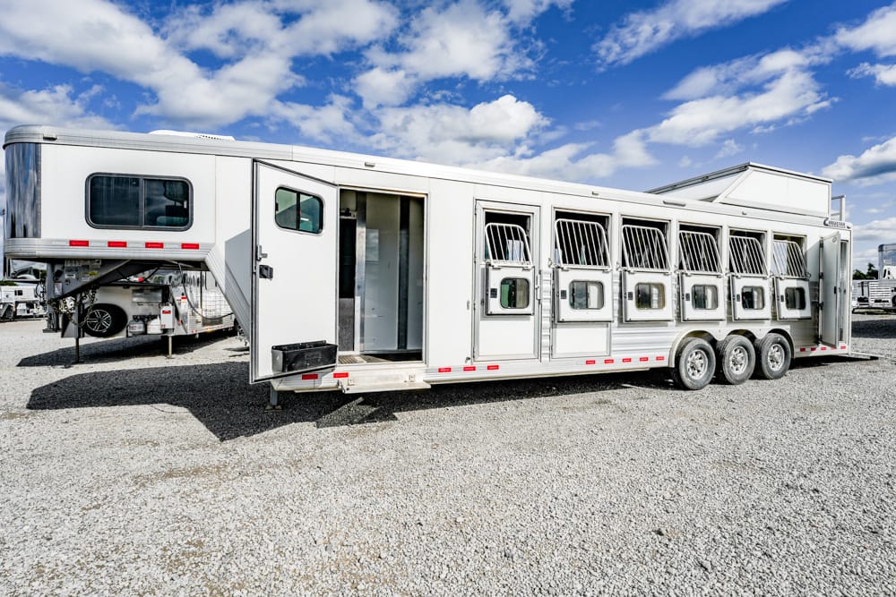 Long white horse trailer with multiple stall doors on a gravel lot under a blue sky; designed to transport horses.