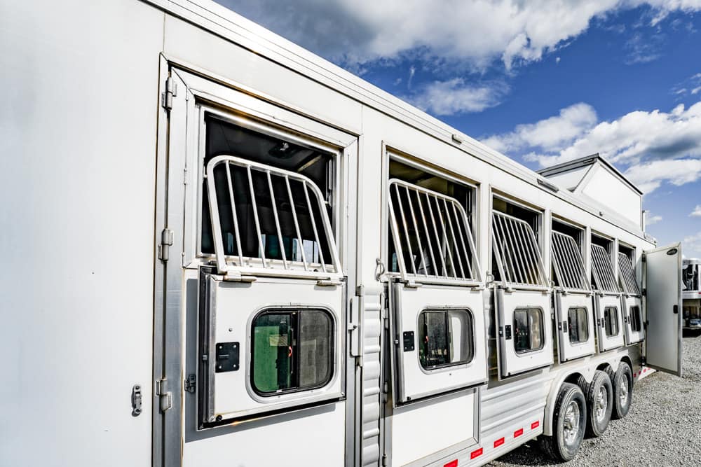White livestock trailer with multiple horse stalls, doors open along the side, parked on gravel under a blue sky.