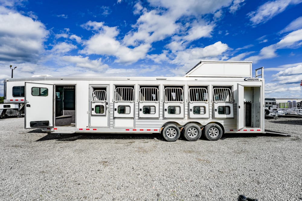 White horse trailer with multiple stall doors and ventilation bars, parked on a gravel lot under a blue sky with clouds.