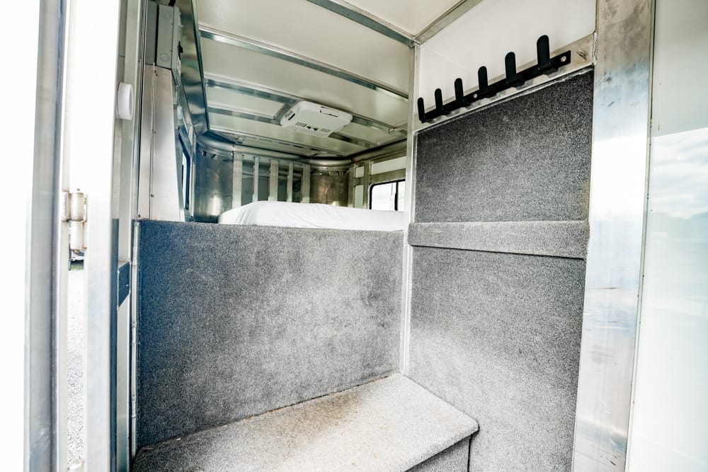 Interior of a cargo trailer with a small bunk on the left, grey carpeted panels, and a row of hooks on the right wall.