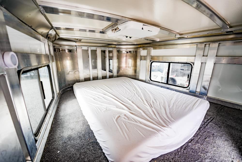 Interior of a metallic trailer converted to a bedroom, with a large white bed on gray carpet and windows along the sides