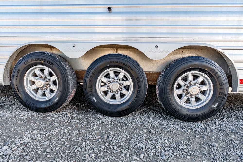 Triple-axle trailer wheels with silver rims on a gravel surface under a corrugated metal trailer.
