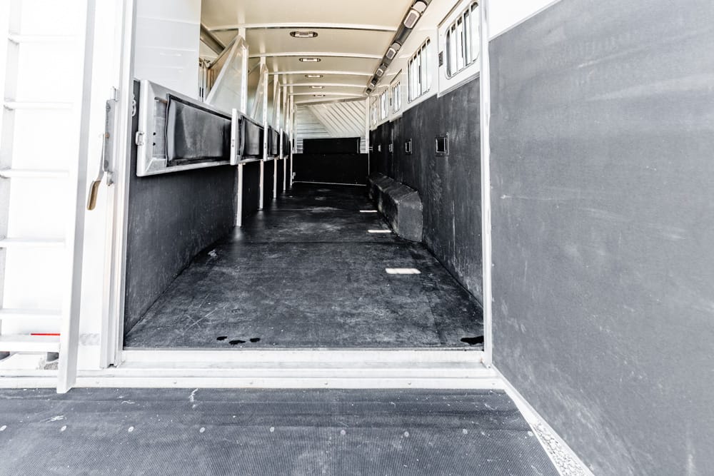 Interior of an empty livestock trailer, dark floor and metal side walls, seen from the doorway with a ramp outside