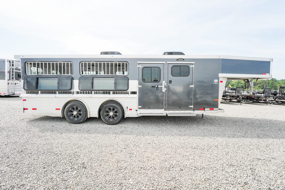Long gray horse trailer with living quarters, two axles and barred windows, parked on a gravel lot on a sunny day