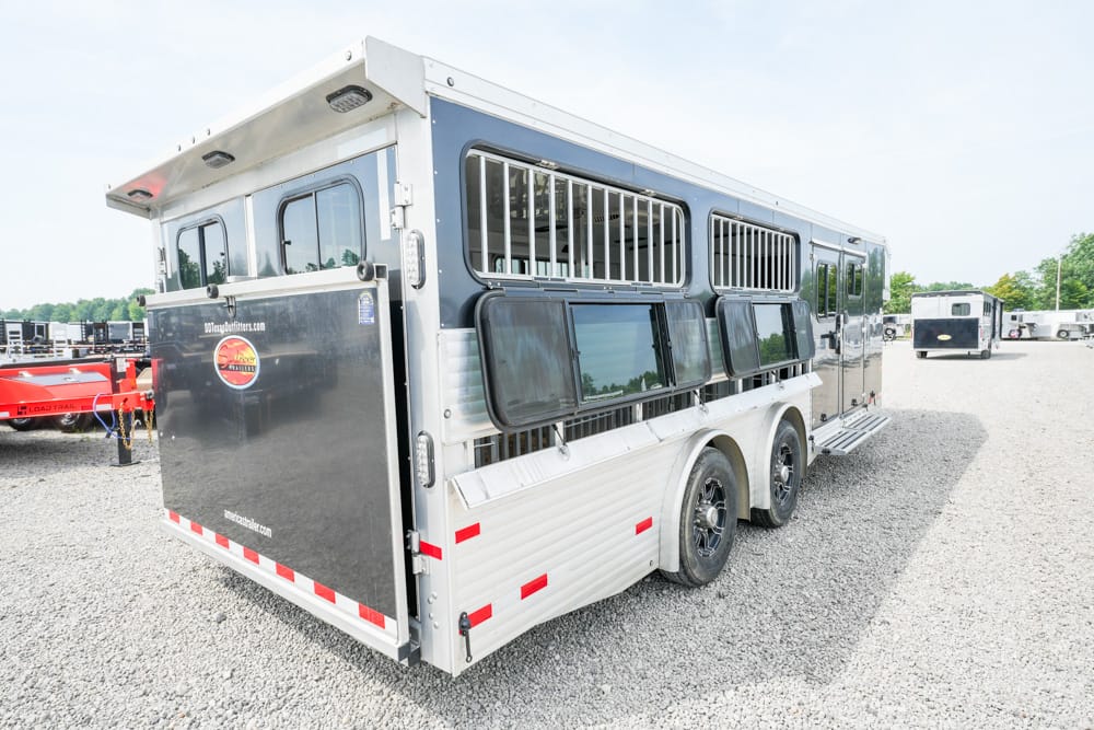 Silver aluminum horse trailer with multiple side windows and vents, parked on a gravel lot with other trailers nearby.