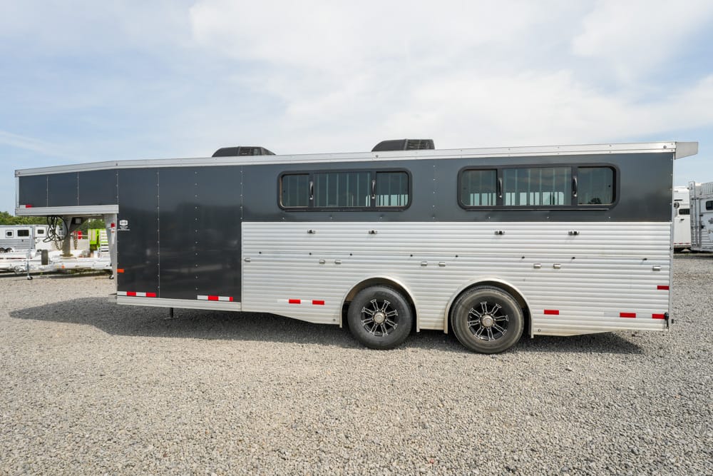 Two-tone horse trailer (dark upper, silver lower) parked on a gravel lot, side view with two windows and dual axles.
