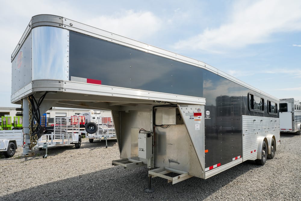 Gooseneck horse trailer with a raised front section, dark blue and silver exterior, parked on a gravel lot with other trailers in the background.