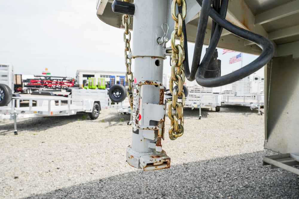 Close-up of a rusted trailer hitch with chains, metal latch, and black hoses in a trailer yard.