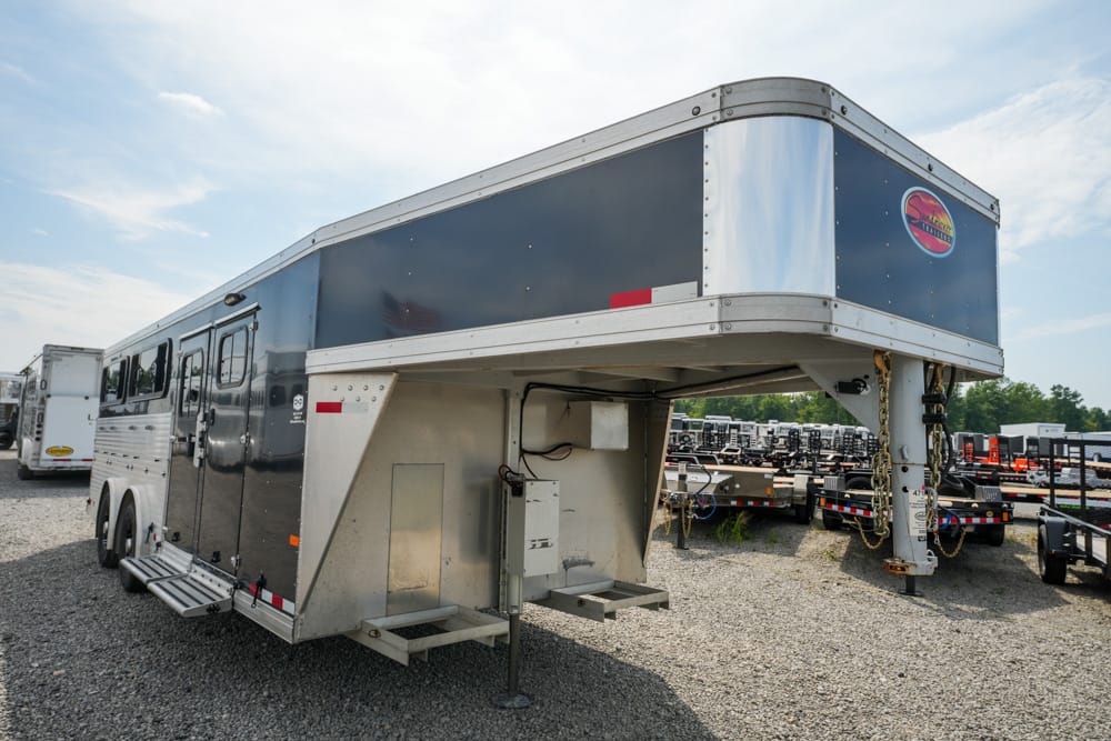 Long blue-gray horse trailer with its gooseneck hitch raised, parked on gravel among other trailers.