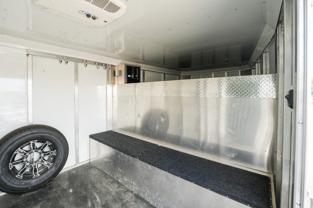 Interior of a aluminum cargo trailer with a long black carpeted ramp/step down the center, a mounted spare tire on the left, and diamond-plate panels along the back wall; ceiling vent visible at top left.