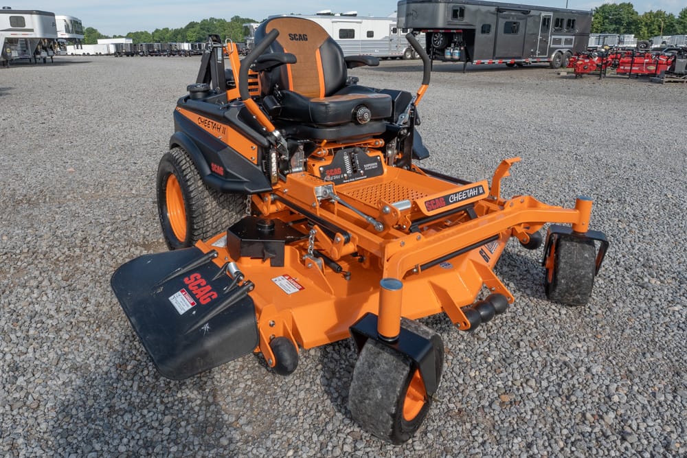 Orange SCAG Cheetah II riding mower with wide cutting deck on a gravel lot, RVs and trailers in the background