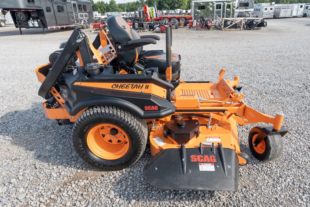 Orange SCAG Cheetah II riding mower with black seat and dual control levers, parked on a gravel lot among equipment trailers and other machinery.