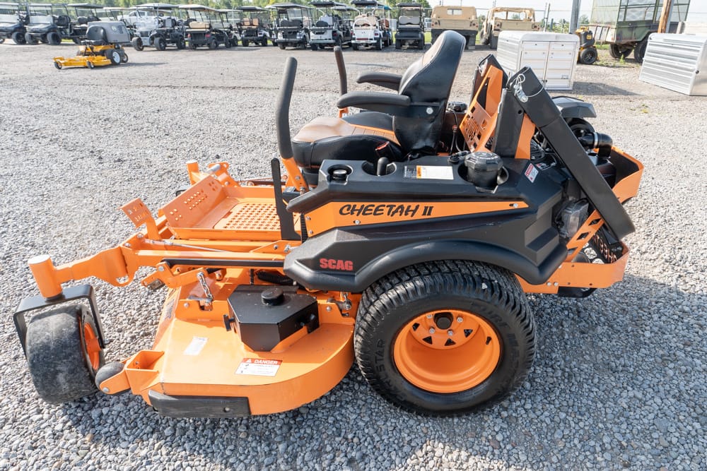 Orange SCAG Cheetah II riding lawn mower on a gravel lot, showing seat, roll bar and cutting deck edge nearby.