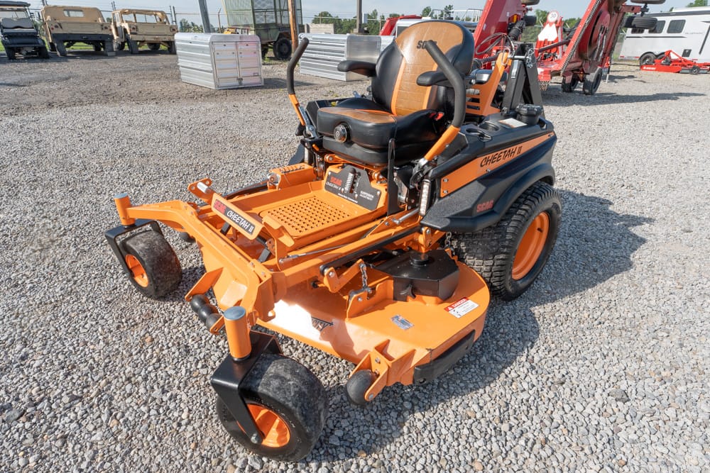Orange Scag Cheetah II zero-turn riding mower with black seat and control levers, parked on a gravel lot among heavy equipment in the background.