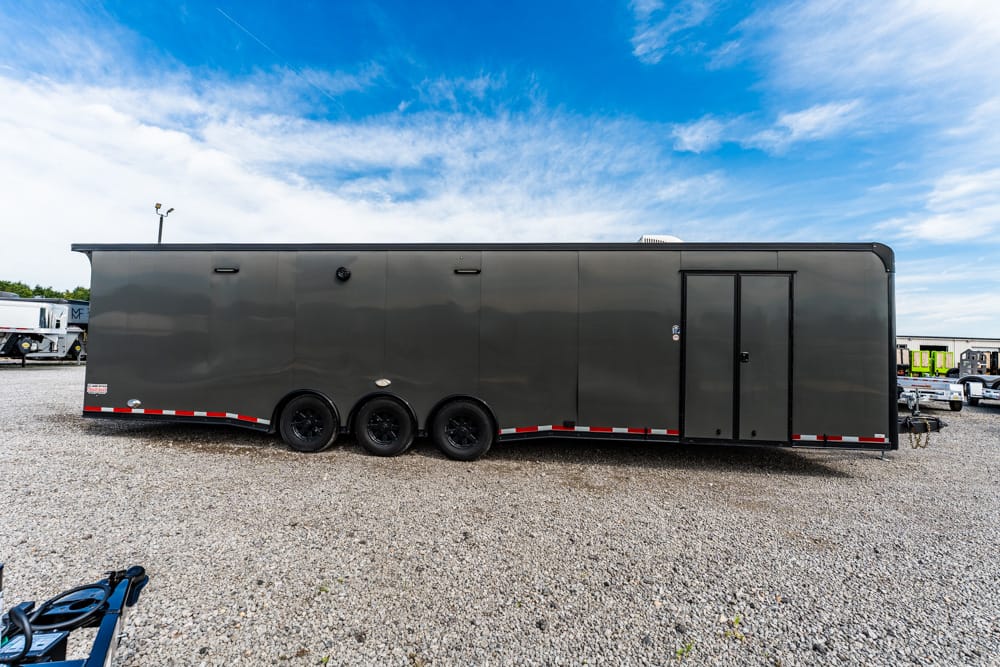 Large dark gray enclosed trailer with three axles parked on a gravel lot under a blue sky with clouds.