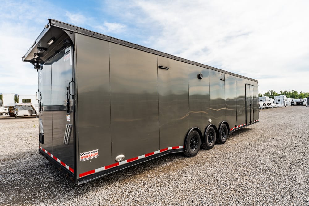 Large gray enclosed trailer with three axles parked on a gravel lot, side door visible and reflective panels.