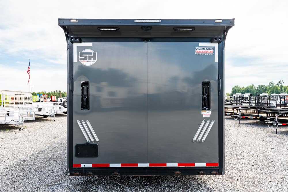 Rear view of a gray trailer with two doors, metal handles, and red-white reflective tape in a gravel trailer yard.