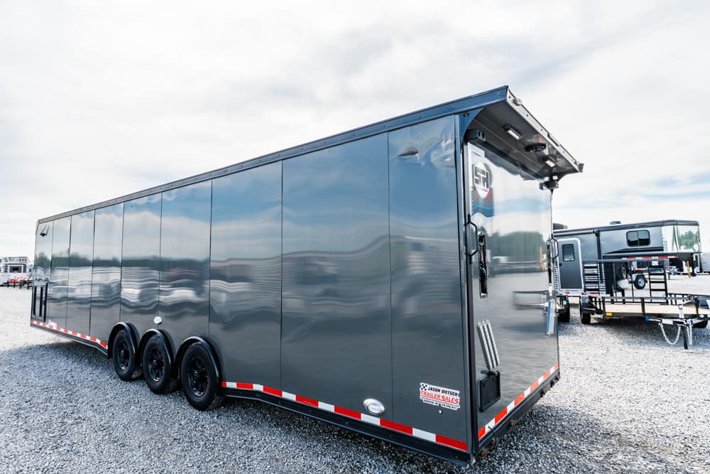 Long gray enclosed trailer with three axles parked on a gravel lot, red-and-white safety stripes along the lower edge.