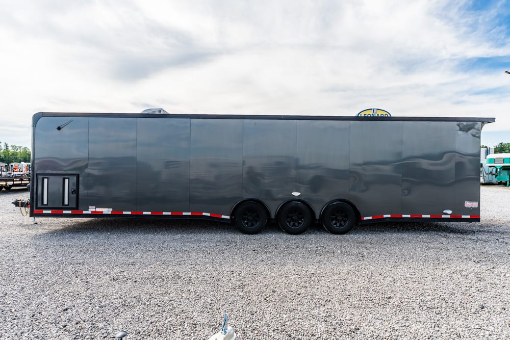 Large gray enclosed cargo trailer with three axles parked on a gravel lot, side profile visible.