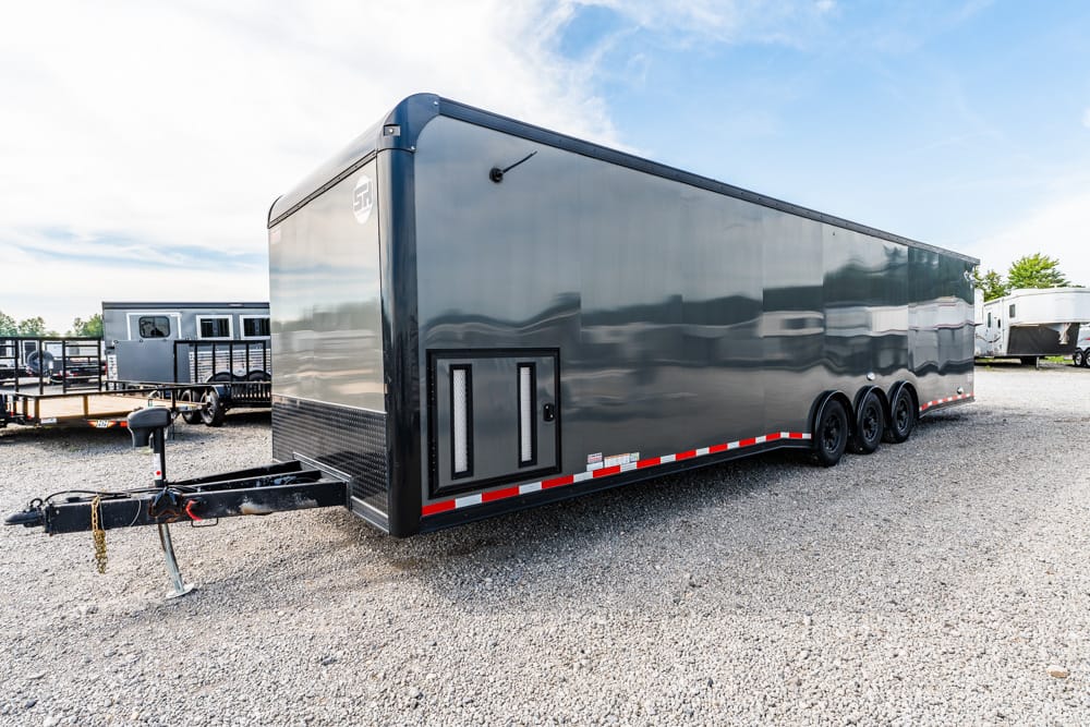 Long glossy gray enclosed cargo trailer with three axles parked on a gravel lot; front hitch and safety chains visible