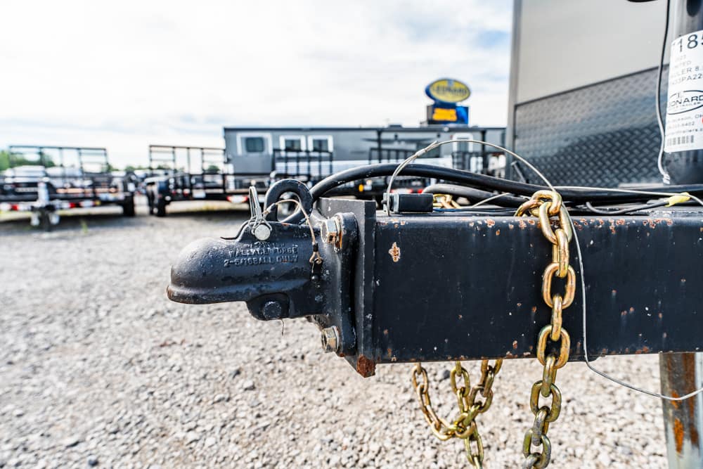 Close-up of a rusted trailer hitch with a thick metal chain, on a gravel lot with other trailers in the background.