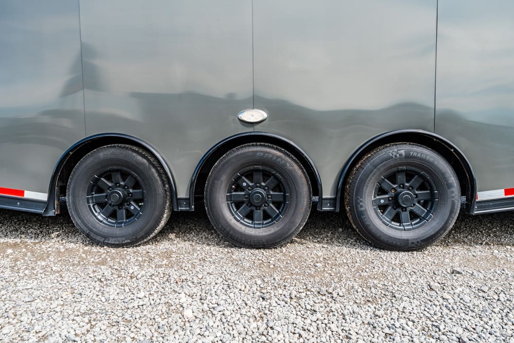 Triple-axle trailer side view with black rims on a gravel surface near wheel wells across the bottom of the trailer FWD edge—on rough ground.