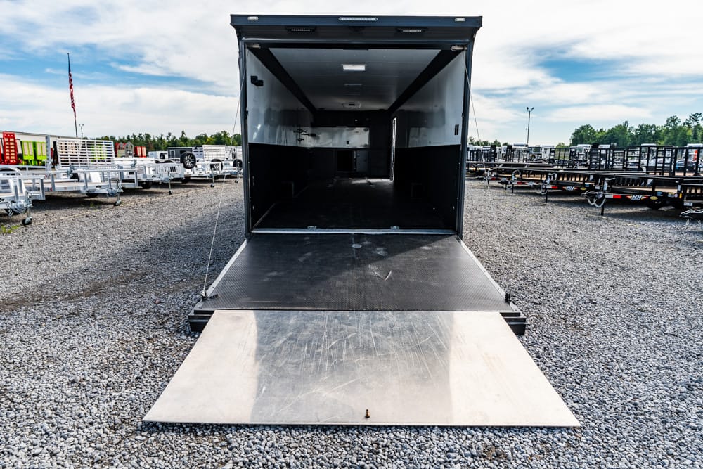 Open black trailer with metal ramp extended onto a gravel lot, rows of trailers in the background.