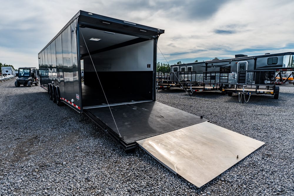 Open black cargo trailer with a lowered rear ramp on a gravel lot, other trailers in the background.