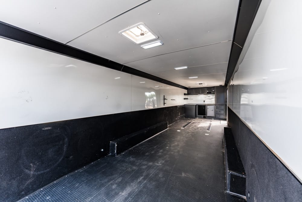 Interior of an empty cargo trailer with white upper walls, black lower panels, and a rubber floor; benches along both sides and storage at the far end.