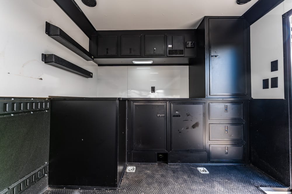 Interior of a black utility kitchen in a trailer: upper cabinets, drawers, and a metal work surface with a diamond-plate floor.