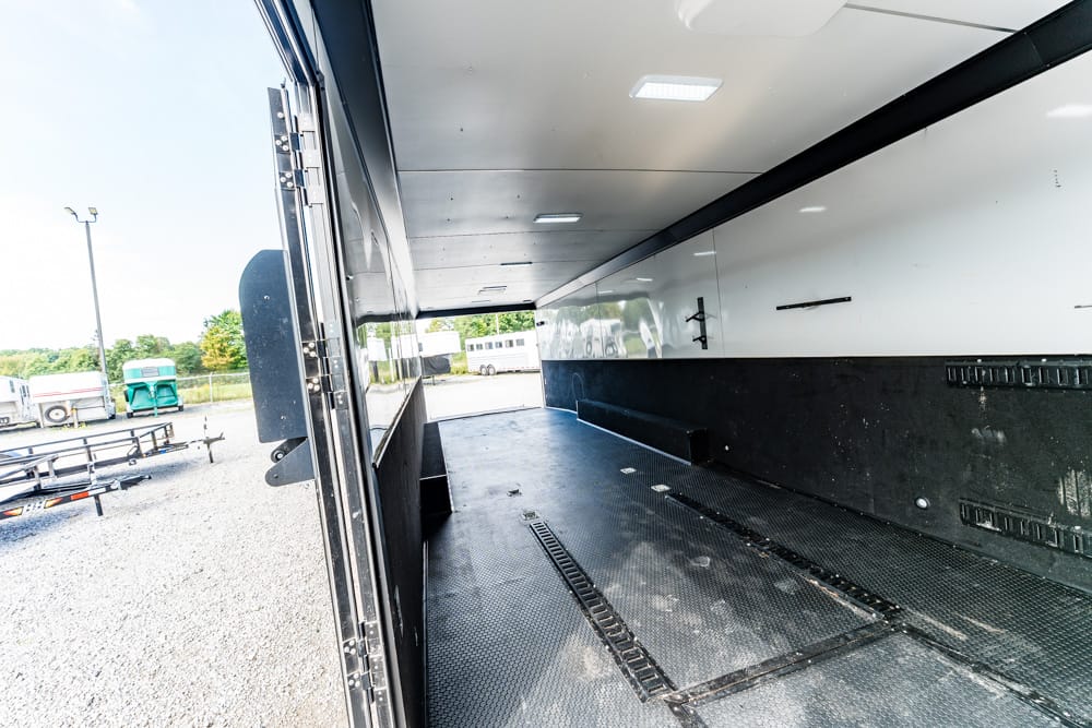 Open cargo trailer interior with black rubber flooring and side walls; parked in a lot with other trailers visible outside the rear opening.
