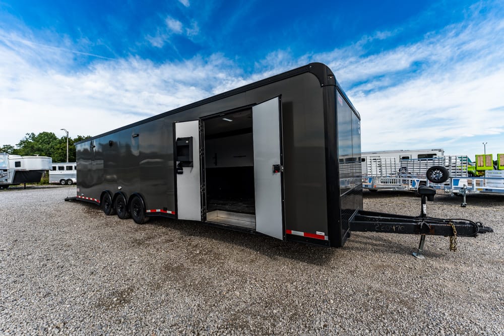 Large black enclosed cargo trailer with open side door, triple axle, in a gravel lot under a blue sky with other trailers nearby.