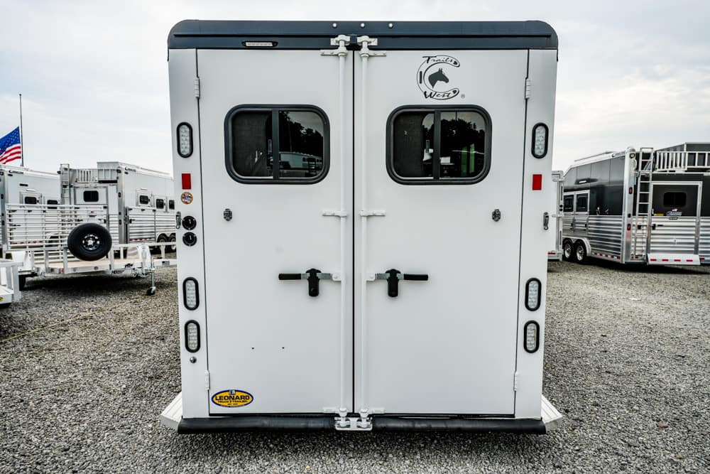 Rear view of a white horse trailer with double doors, small windows, and black latches on a gravel lot with other trailers nearby.