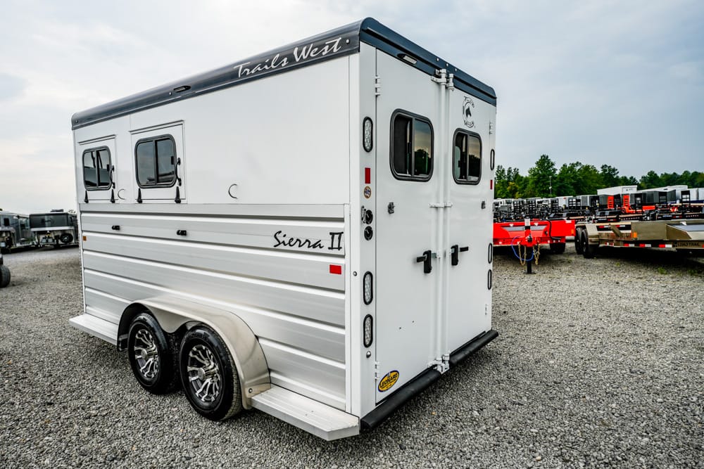 White tandem-axle horse trailer with 'Trails West' on the top edge and 'Sierra II' on the side, parked on a gravel lot with other trailers in the background