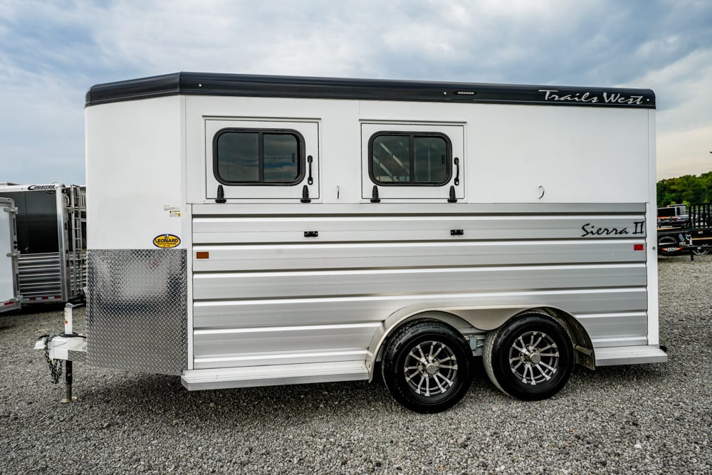 White horse trailer with two side windows and dual axles on a gravel lot.