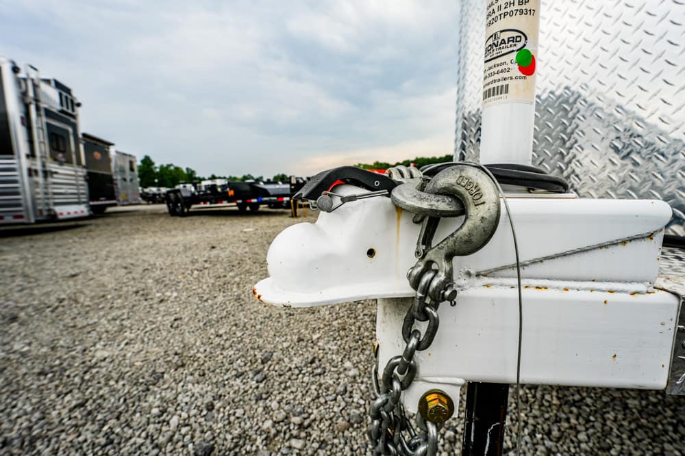 White trailer hitch with a heavy metal lock and chain secured to a white trailer in a gravel lot with other trailers in the background.