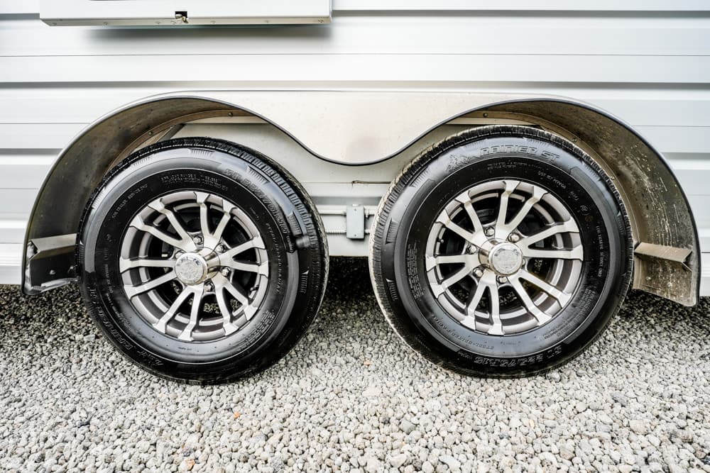 Twin trailer wheels with silver rims on a gravel ground beside a white trailer body.