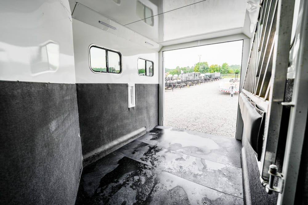 Interior of an empty white trailer with black padded walls and small windows, open to a gravel lot showing other trailers outside in daylight.