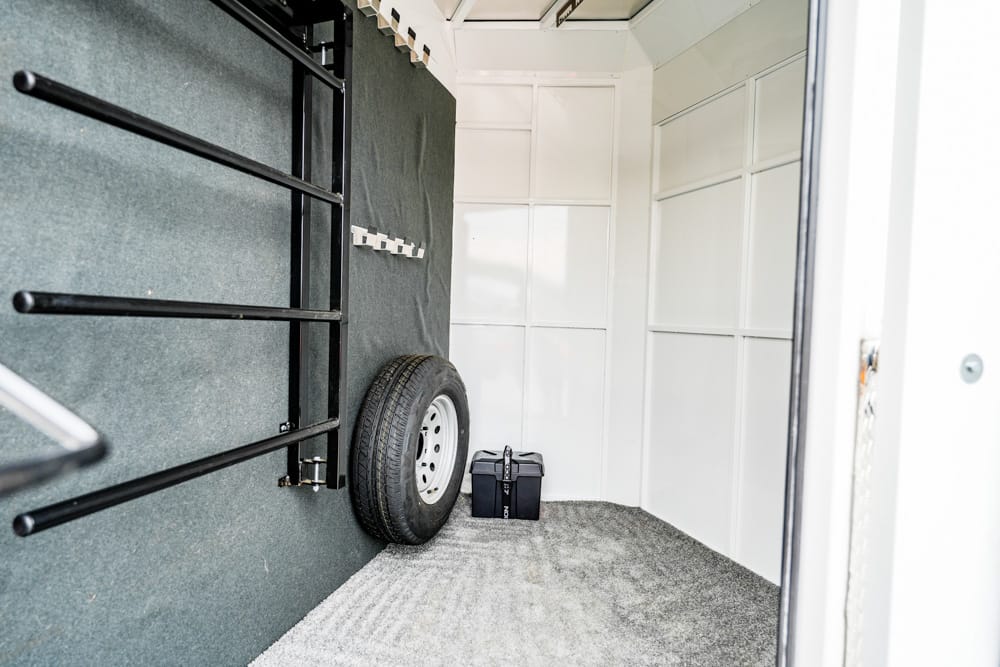 Interior of a utility trailer: gray carpeted floor, white walls, spare tire on the left wall, and a black toolbox on the floor near the rear door with wall-mounted racks nearby.
