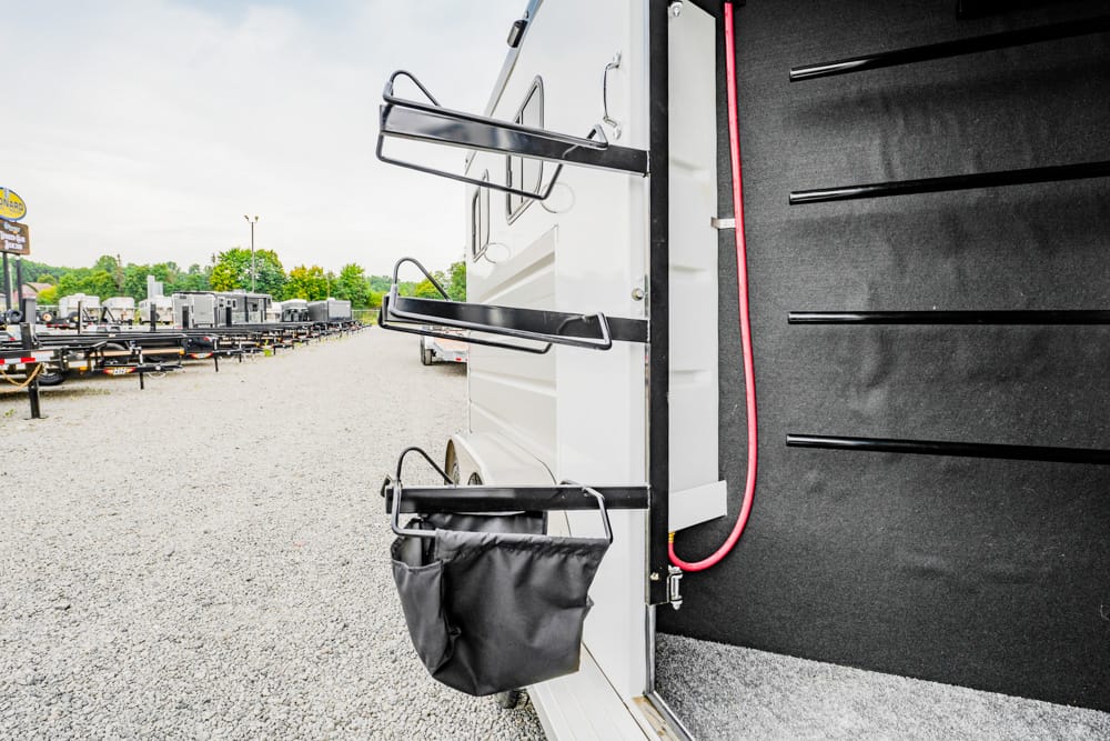 Back of a white trailer with black storage racks and a hanging black bag; a red power cord runs along the doorway, on a gravel lot with other trailers in the background.