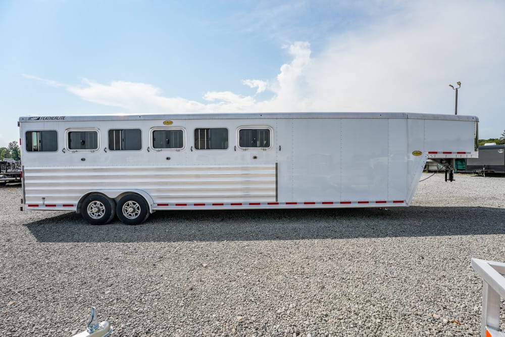 White aluminum horse trailer with multiple windows along the side, two axles, parked on a gravel lot under a blue sky.