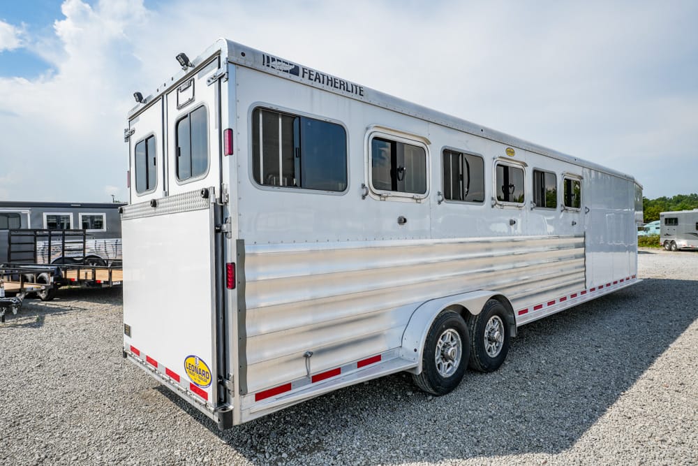 White Featherlite horse trailer with multiple side windows and dual axles on a gravel lot, viewed from rear-right corner.