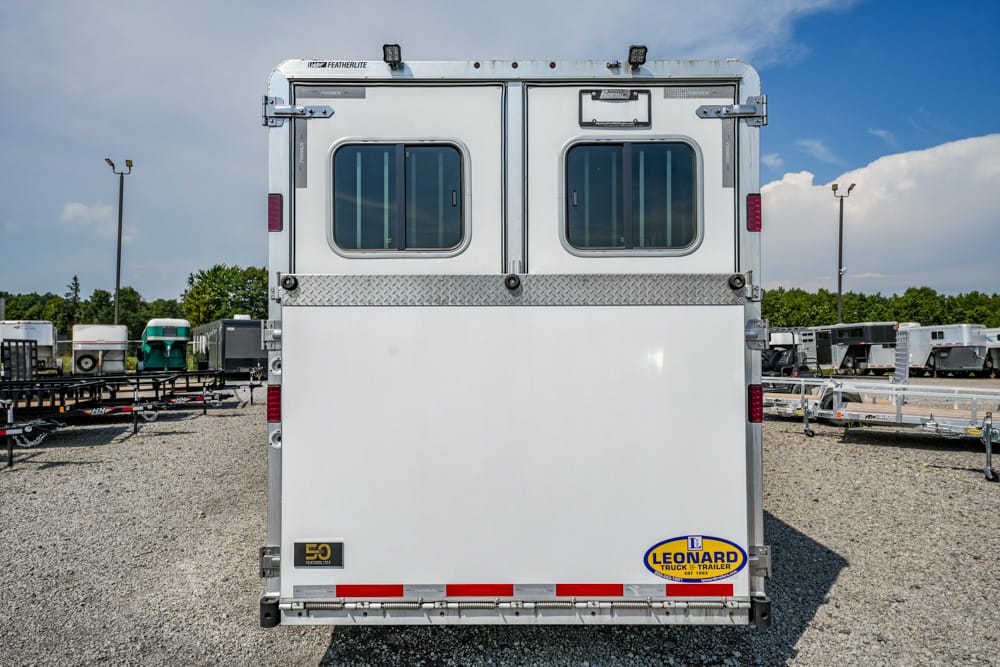 Rear view of a white double-compartment horse trailer with two small windows, diamond-plate trim, and a Leonard trailer logo on a gravel lot.