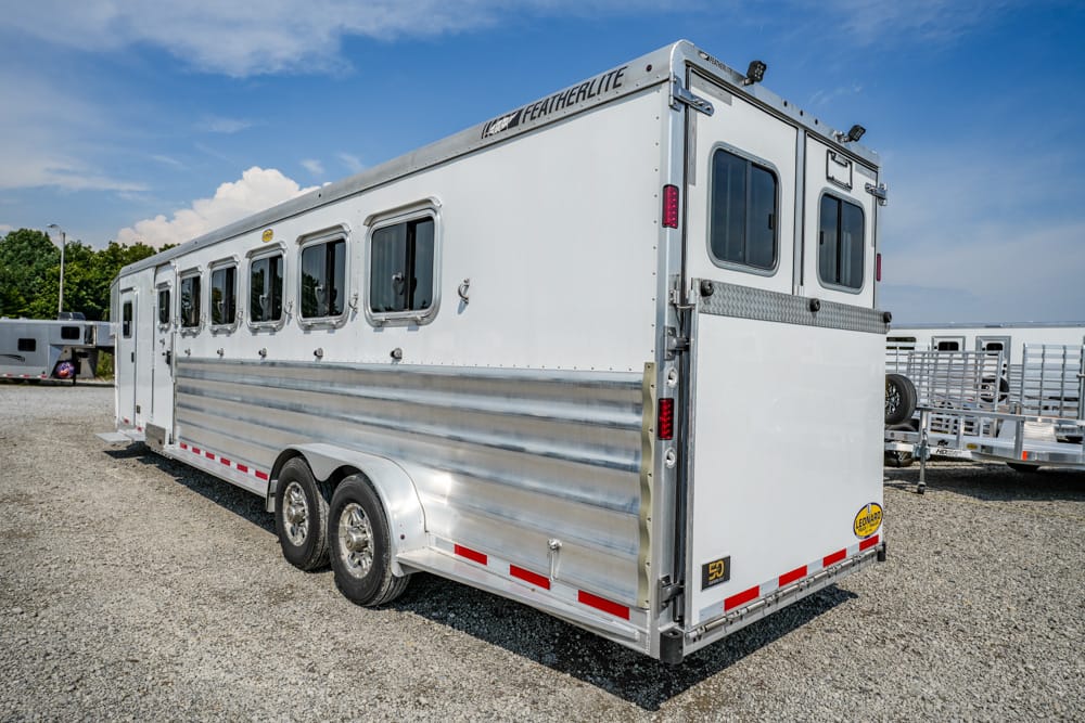 White aluminum horse trailer with multiple windows and dual axles on a gravel lot under a blue sky.