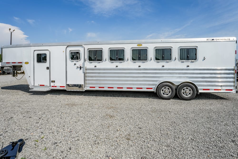 Long white aluminum horse trailer with multiple windows, parked on a gravel lot under a clear blue sky.